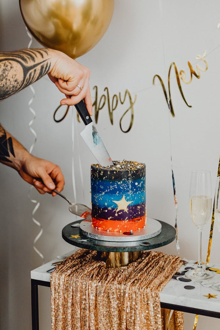A tattooed hand slices a vibrant cake at a festive New Year celebration.